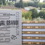 Libro ¡Al Grano! de Kim Ossenblok de pie junto a una pila de nueve ejemplares, con casas y árboles de un pueblo al fondo: regalo para amantes del café