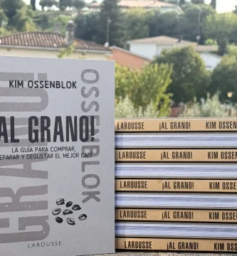 Libro ¡Al Grano! de Kim Ossenblok de pie junto a una pila de nueve ejemplares, con casas y árboles de un pueblo al fondo: regalo para amantes del café